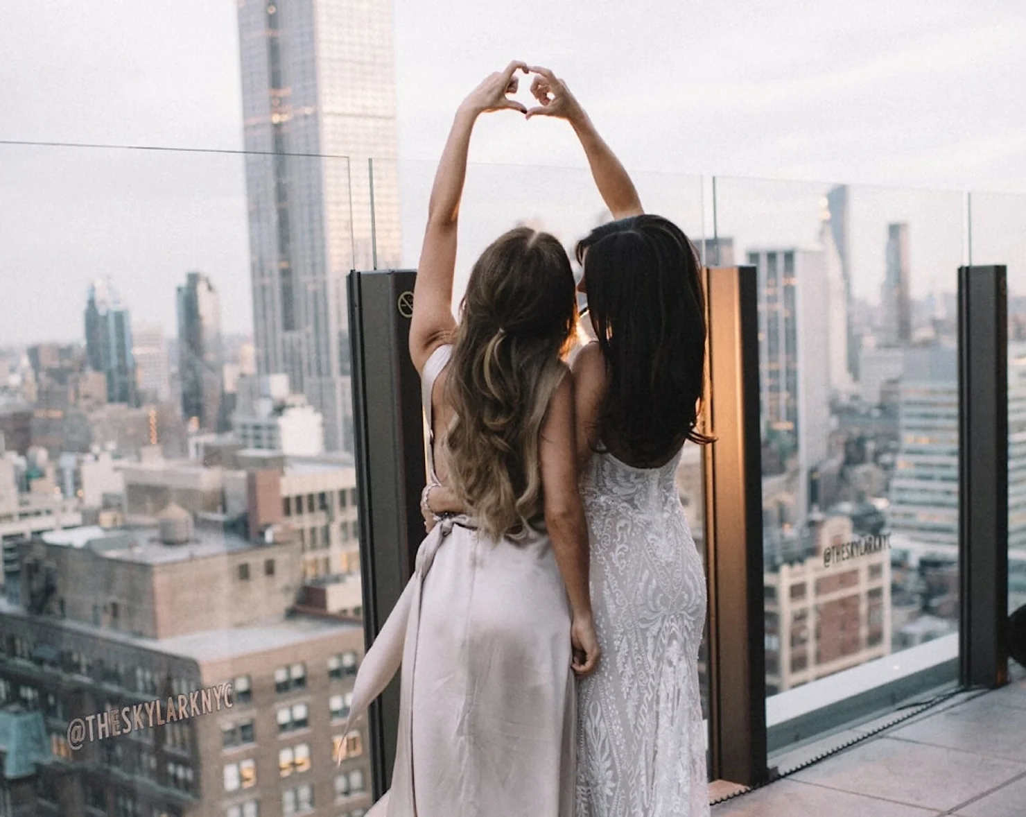 nyc wedding dj skylark wedding Two women in white dresses on a rooftop form a heart with raised hands, city skyline behind, @THESKYLARKNYC visible.