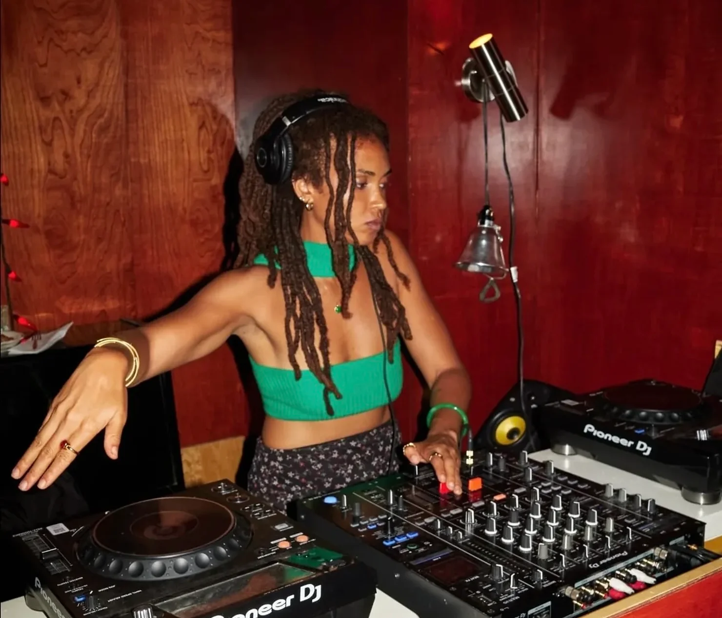 nyc wedding dj new york wedding timeline Femaile Jamaican DJ in headphones mixes on a Pioneer console in a wood-paneled booth, wearing a green top, focused and immersed.