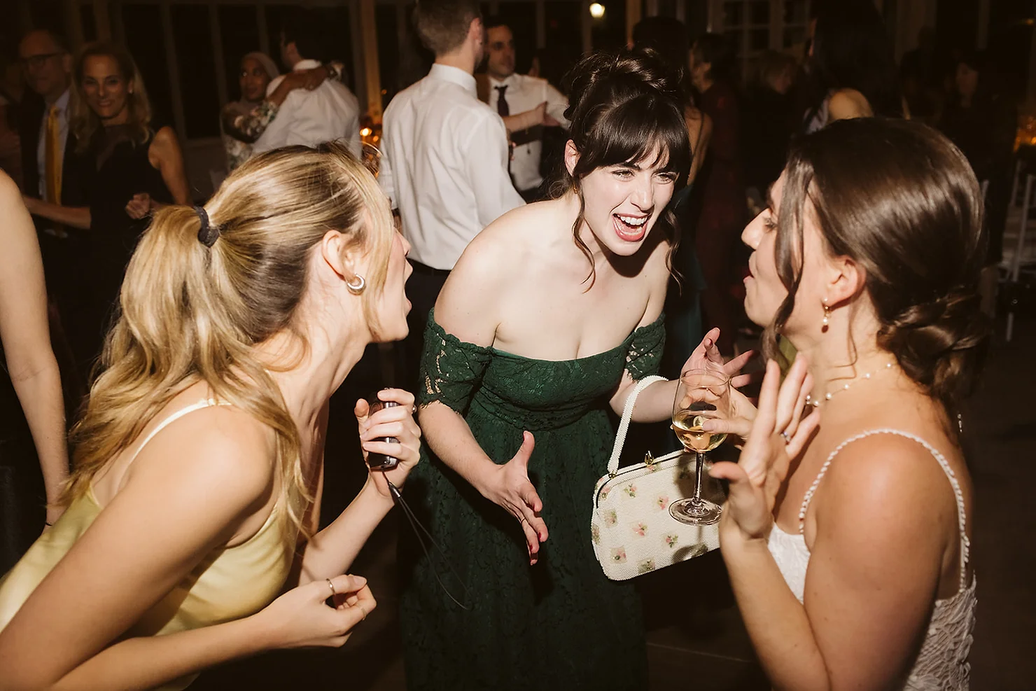 NYC Wedding DJ Women laughing and chatting at a dimly lit party, one in a green dress holding a wine glass and floral purse.