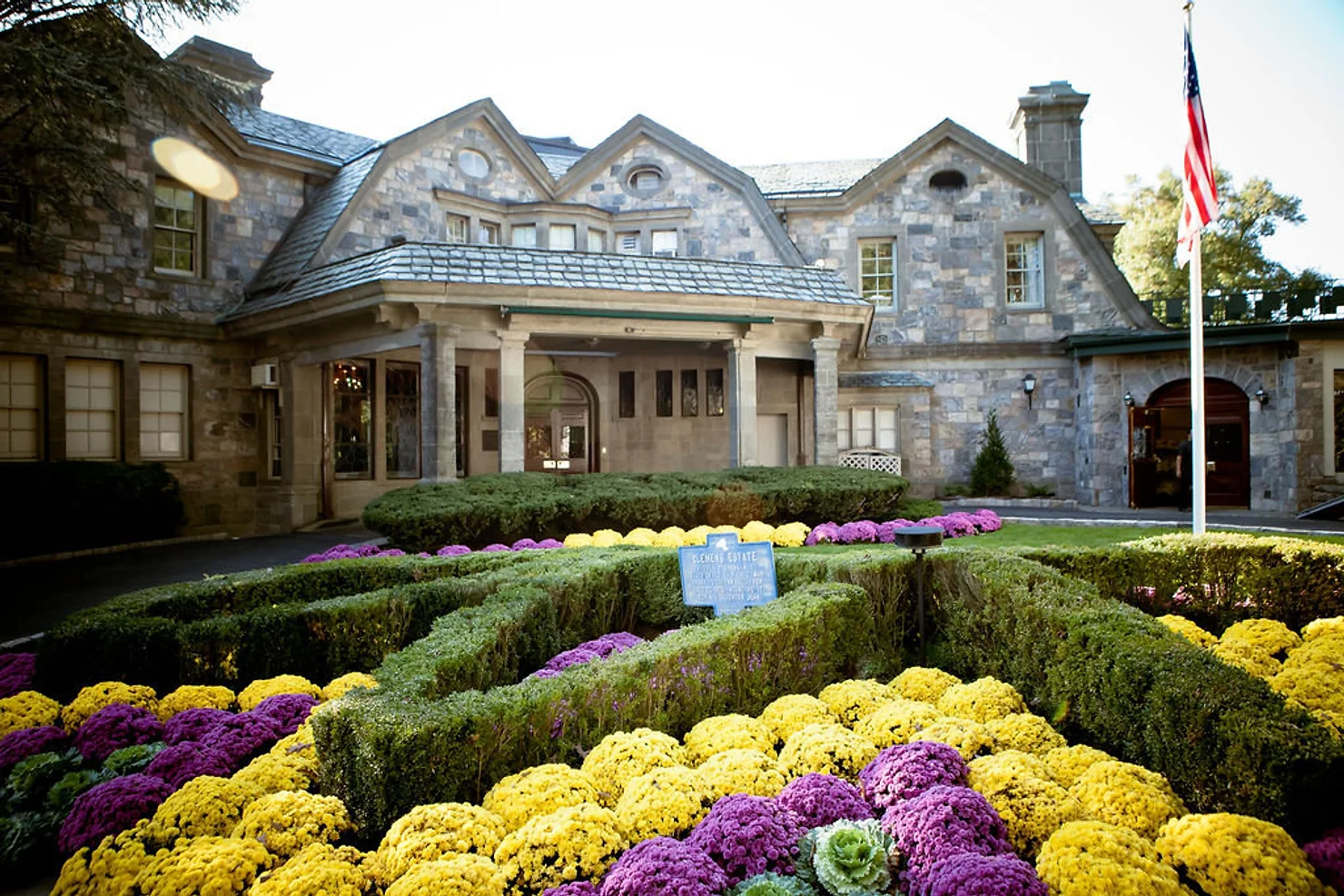 nyc Tappan Hill Mansion by Abigail Kirsch non traditional wedding dj: Stone mansion behind manicured hedge maze and yellow-purple flowers, with an American flag flying outside.