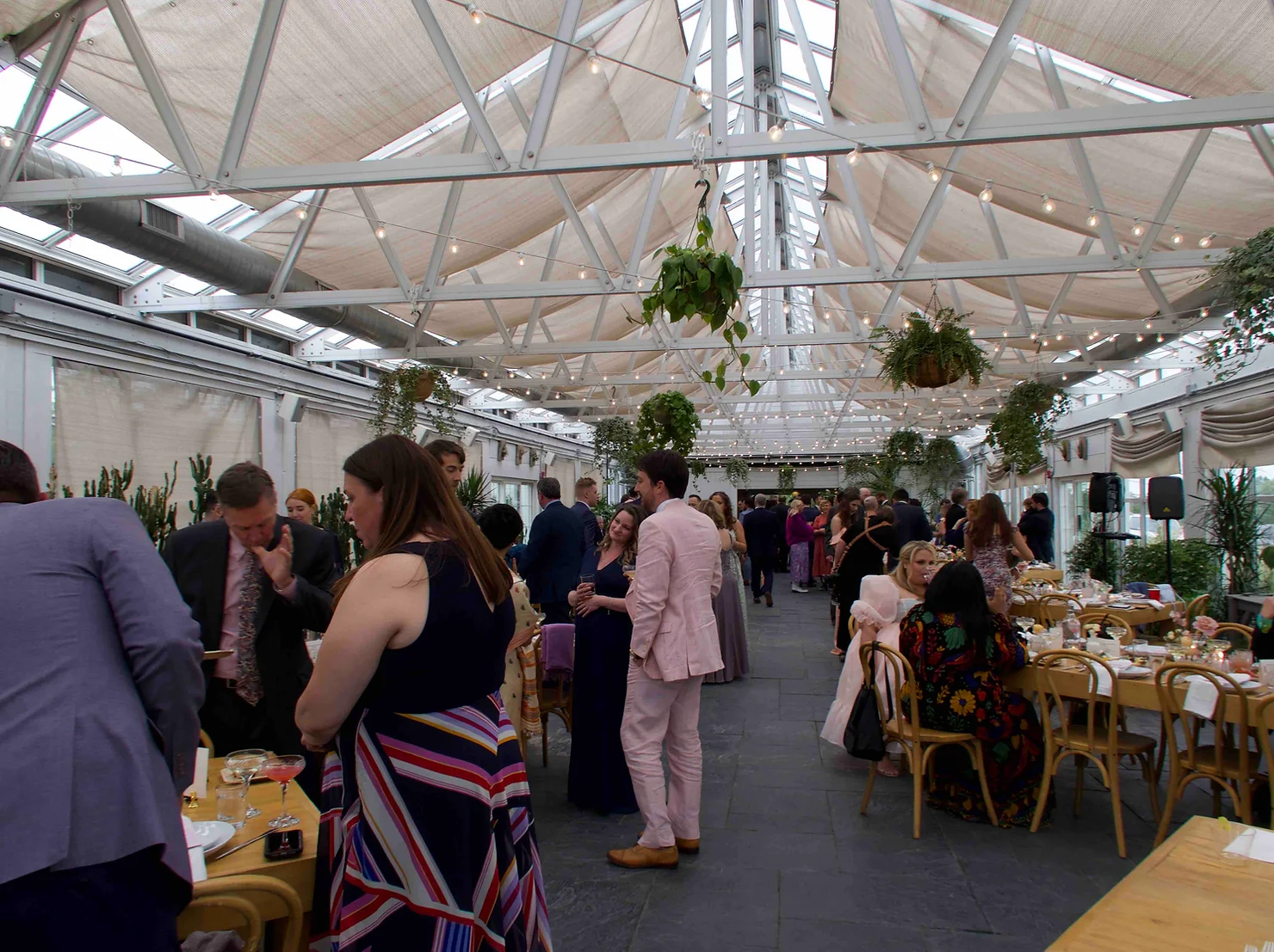 nyc wedding dj Guests mingle at a lively indoor rooftop reception under skylights and hanging plants, with long dinner tables and string lights.
