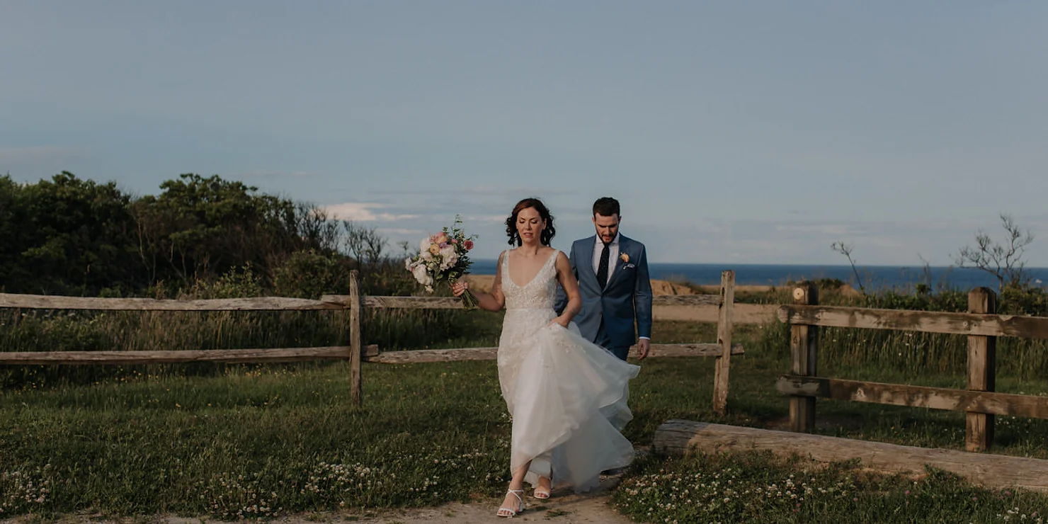 Wedding at the Montauk. Bride in white dress holding flowers leads groom in blue suit along a rustic fence path. Ocean in background under blue sky.