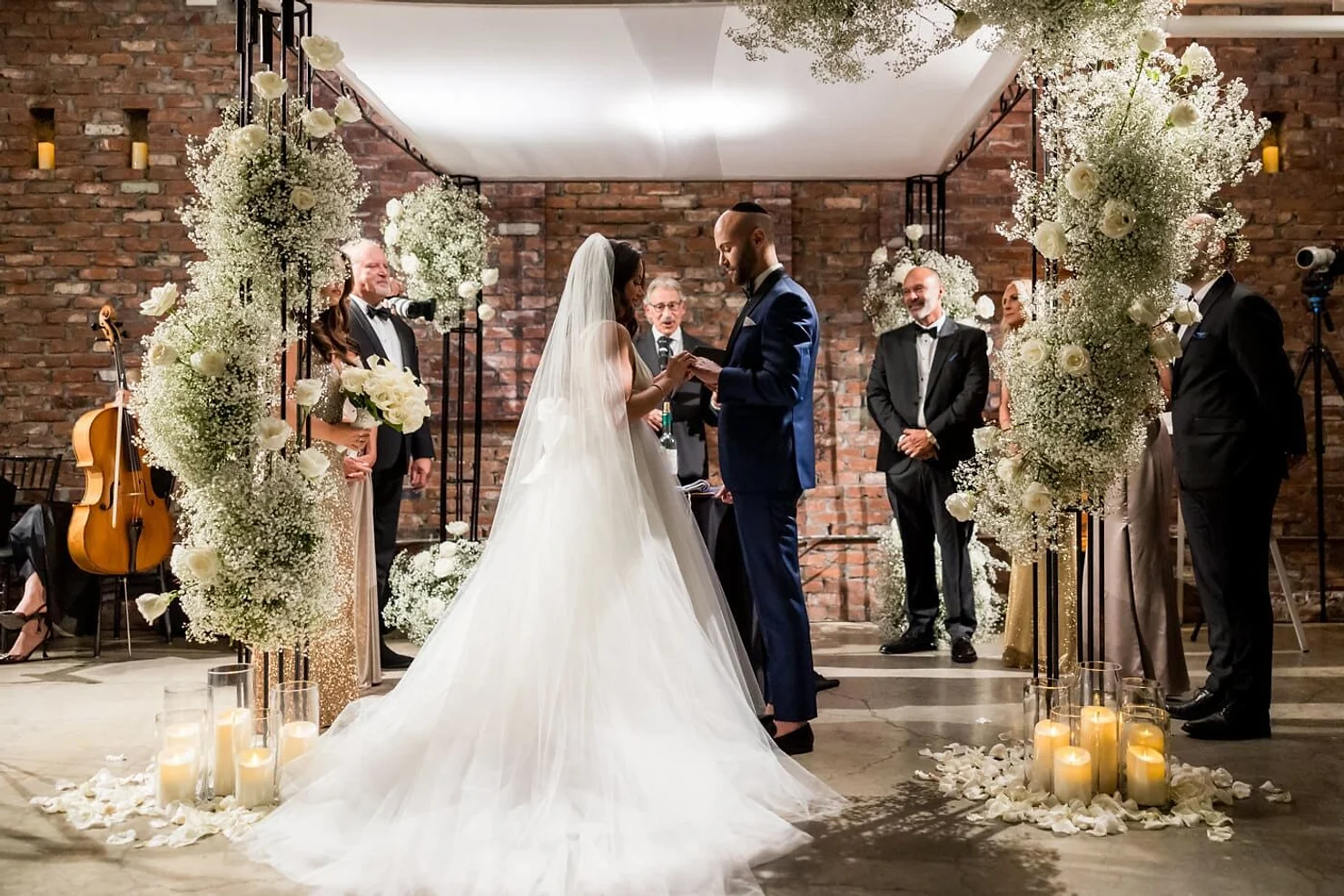 The Loading dock non traditional wedding DJ. Bride and groom exchange rings at a candlelit wedding ceremony, framed by white floral arches and guests in formalwear.