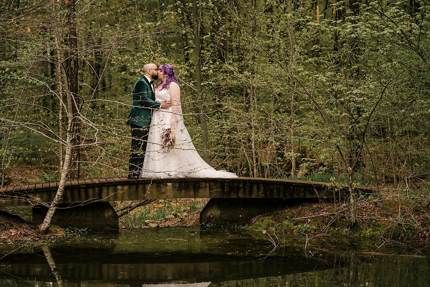 Foxfire Mountain House Wedding DJ: Bride with purple hair kisses groom in dark green suit on a wooden bridge in a lush forest pond scene.