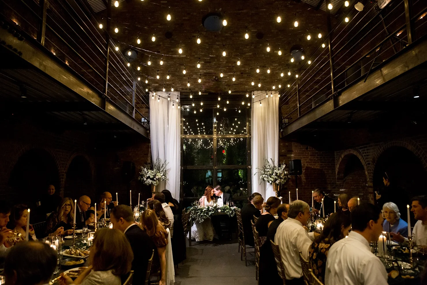 The Foundry nyc non traditional wedding DJ Newlyweds kiss at a candlelit wedding reception in a brick loft, with guests dining under warm string lights.