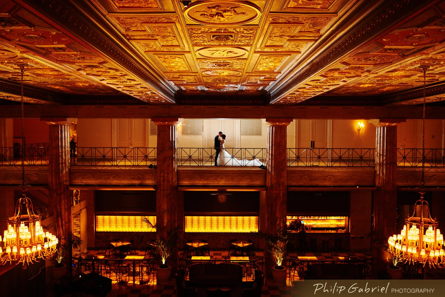 Philly wedding dj Couple kissing on an upper balcony in an ornate golden hall with chandeliers, warm amber lighting, and a grand painted ceiling.