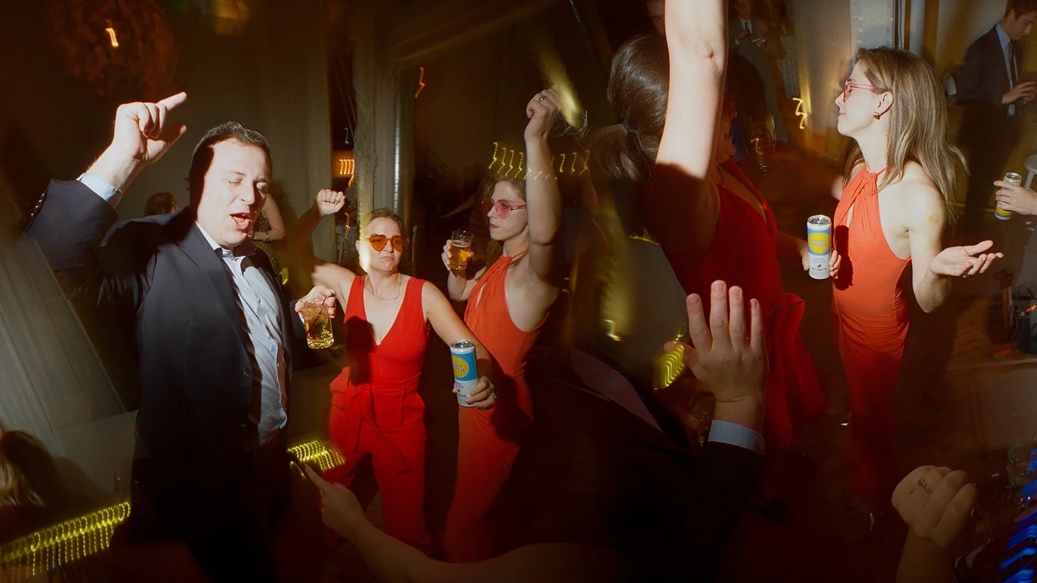 Philly wedding dj People dancing at a dim party, a man in a suit and women in red dresses raising drinks under warm club lights.