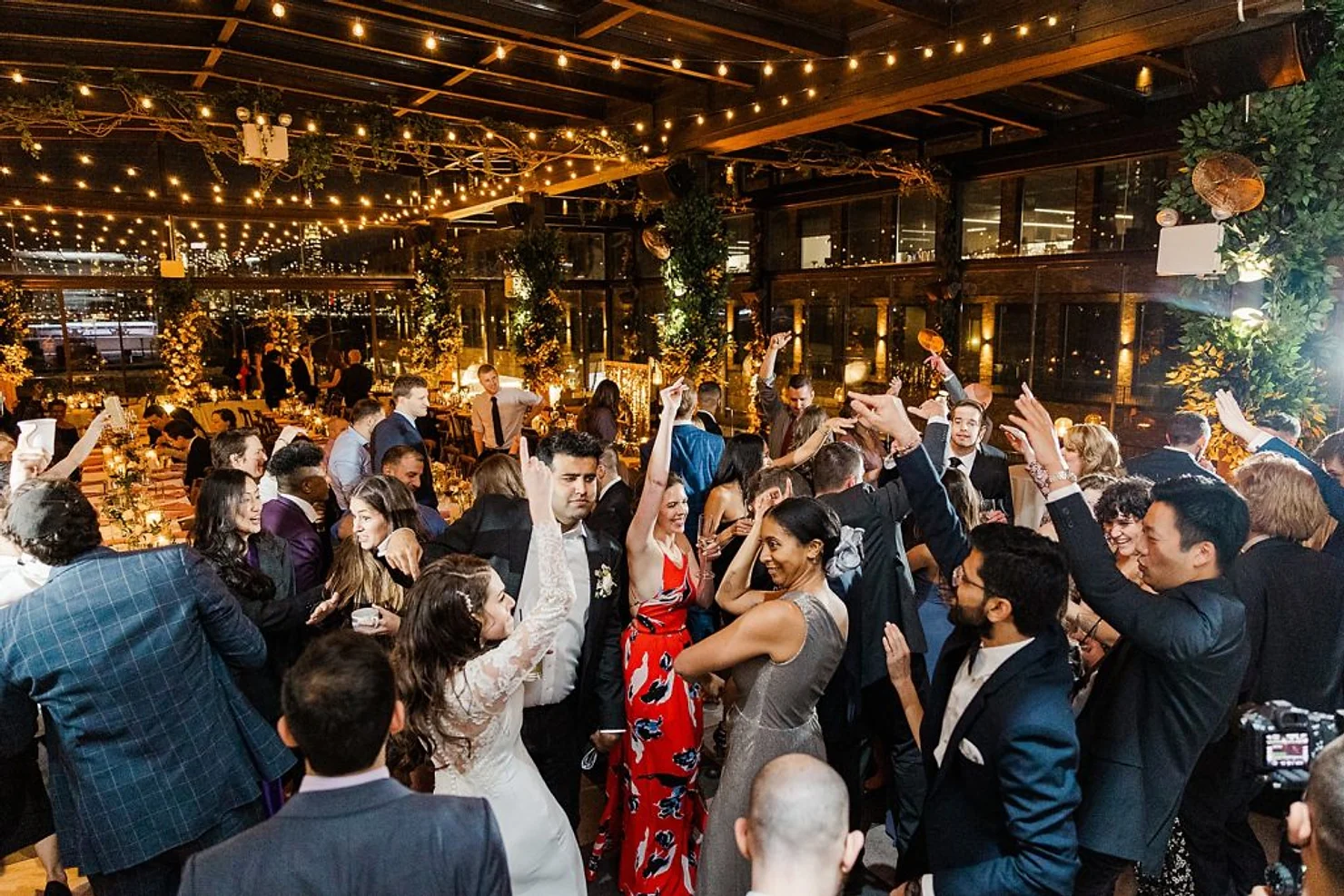 nyc 74 wythe wedding dj  Wedding guests dance under string lights in a warm indoor venue, with bride in white and a woman in red floral dress smiling.