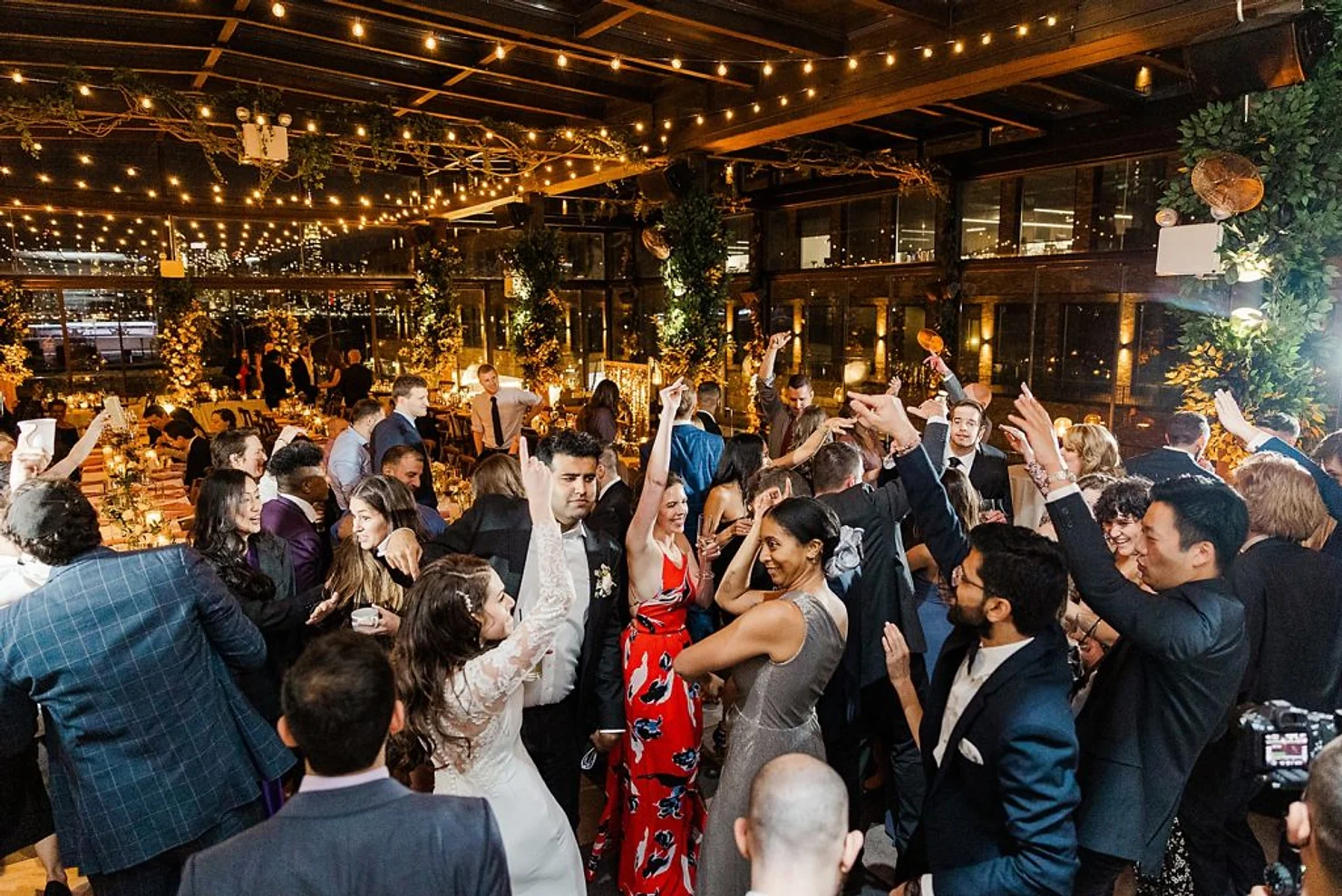 nyc 74 wythe wedding dj Wedding guests dance and cheer in a warm-lit reception hall under string lights, dressed in formal attire.