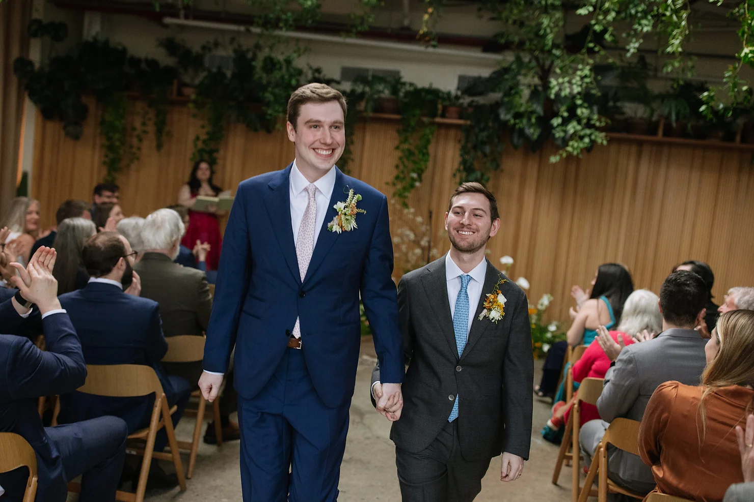 How NYC Wedding DJs Approach Fairfield County Weddings Differently. Two smiling grooms in suits walk down a wedding aisle hand in hand as guests applaud in a greenery-filled indoor venue