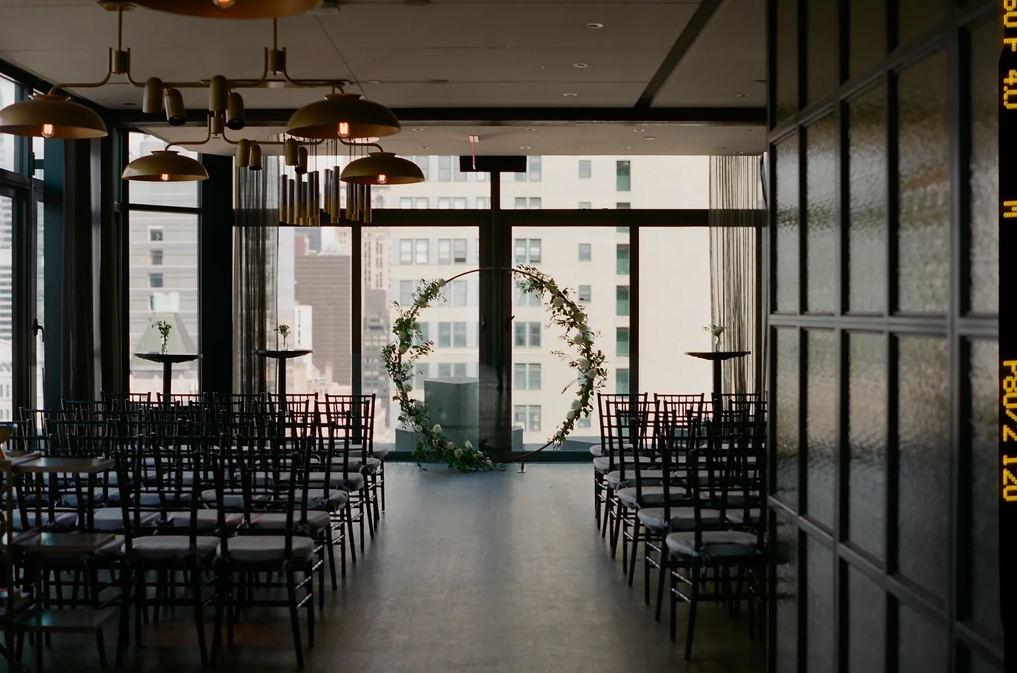 nyc wedding dj skylark wedding Empty modern event hall with rows of black chairs, floral arch by windows, and warm pendant lights overlooking city buildings.