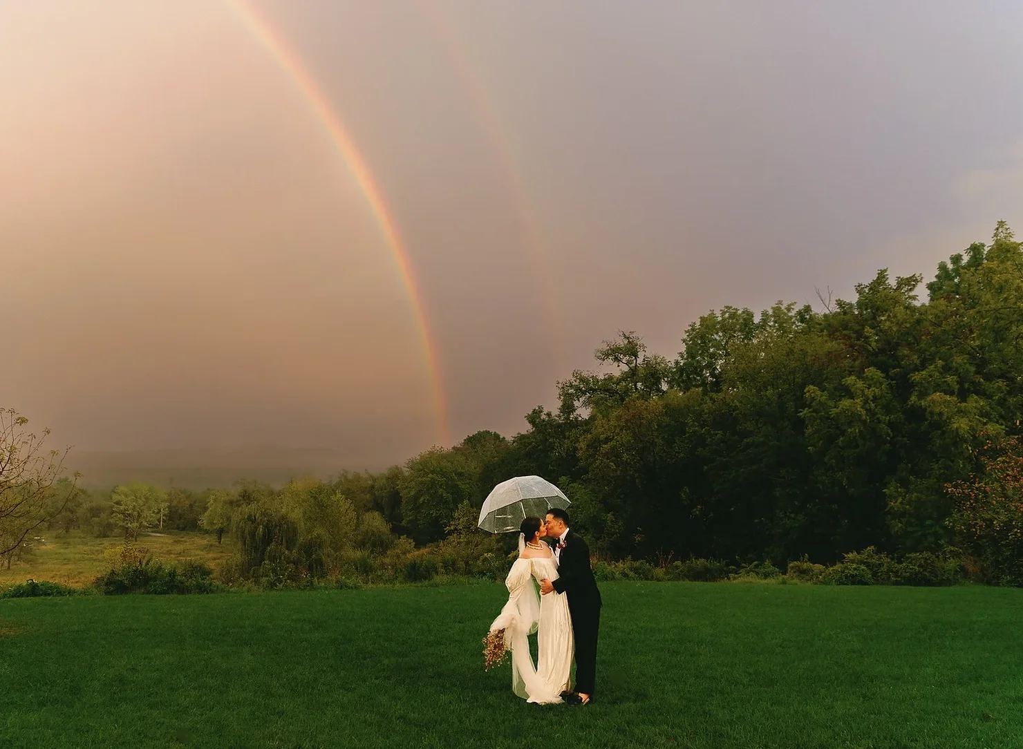 Wedding couple kissing under a clear umbrella on a green lawn, with a rainbow and trees after rain.
