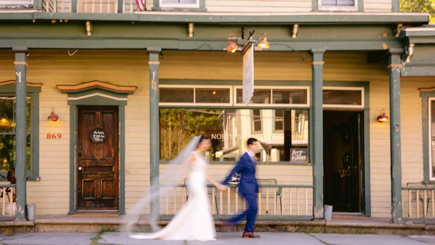 nyc catskills wedding dj North Branch Inn Blurred bride and groom walk hand in hand past a vintage beige storefront with green trim and 869, evoking a wedding moment.