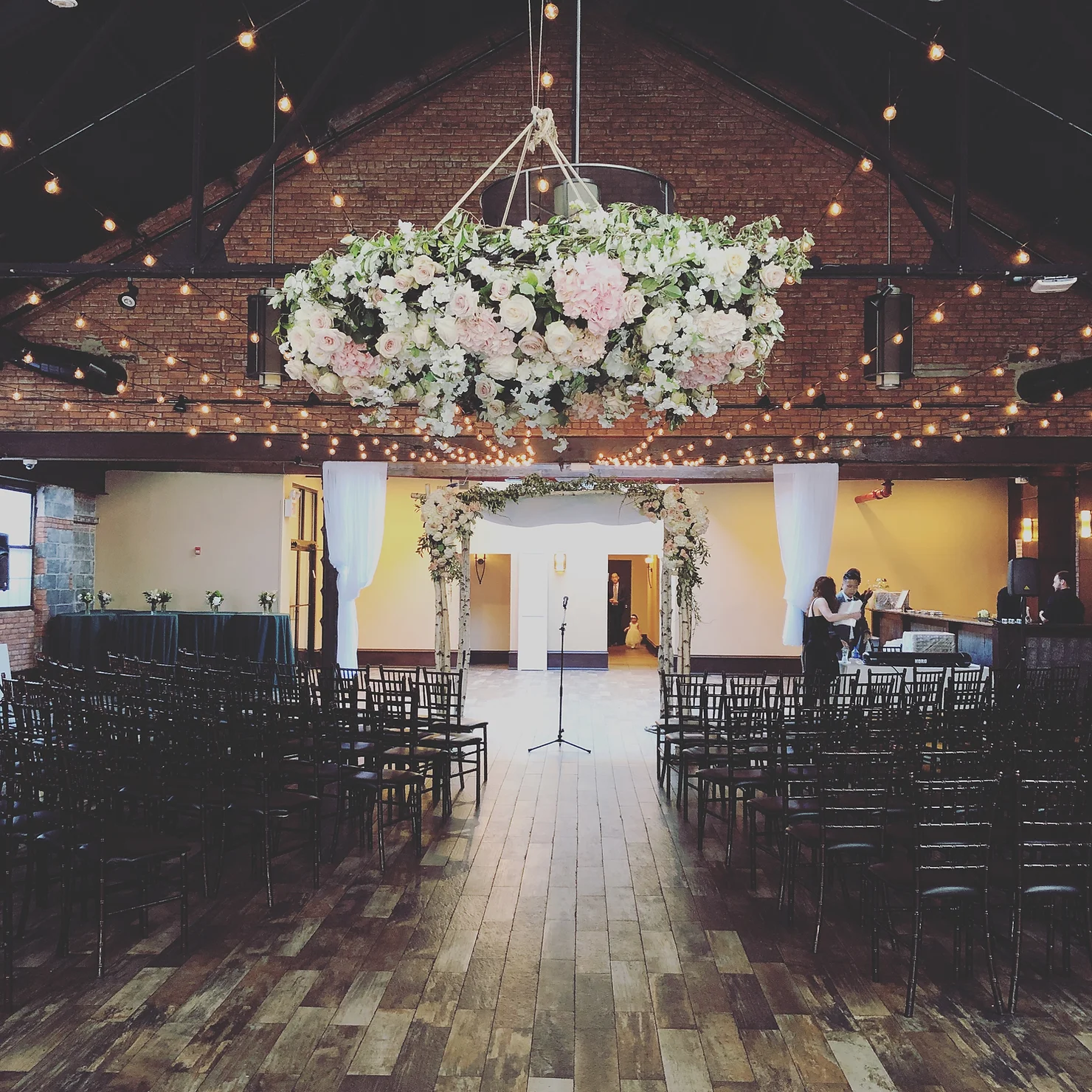 Flower-covered wedding arch in rustic brick hall with string lights, empty rows of black chairs, and a microphone at center aisle