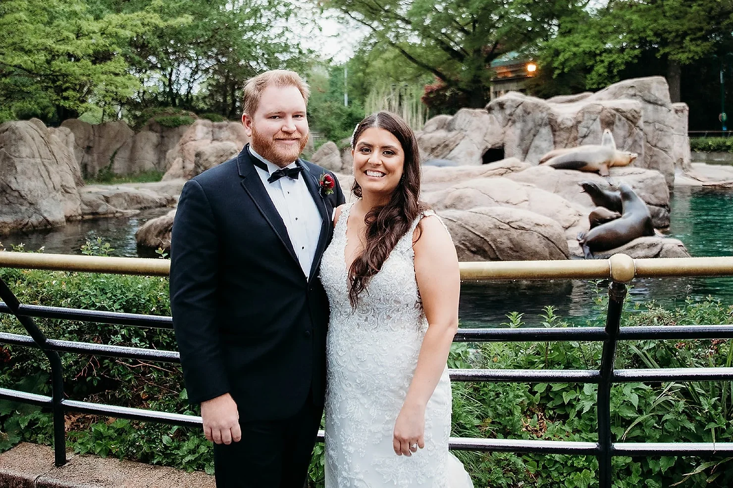 nyc non traditional wedding dj Smiling bride and groom in wedding attire pose by sea lions and rocks at a zoo, with lush green trees behind them.