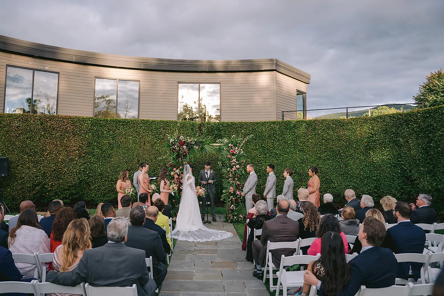 nyc wedding dj foxfire mountain house Outdoor wedding ceremony with bride, groom, bridal party, and guests seated before a floral arch and green hedge under cloudy skies