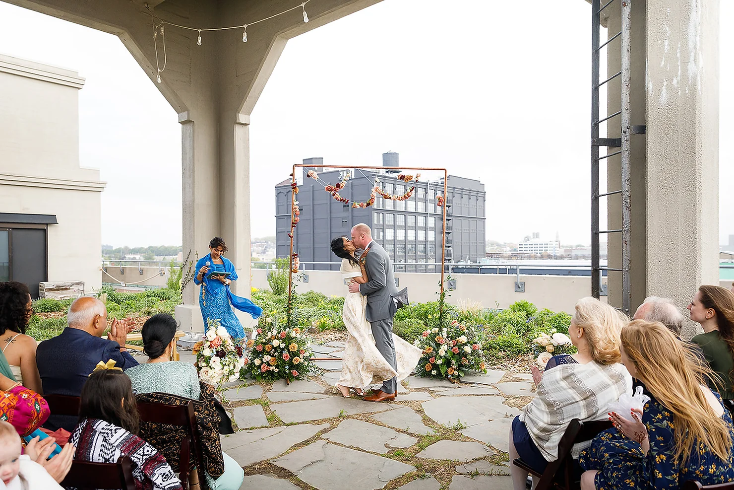 nyc wedding dj new york wedding timeline Bride and groom kiss at a rooftop wedding ceremony, guests applauding under a floral arch with city buildings in the background.