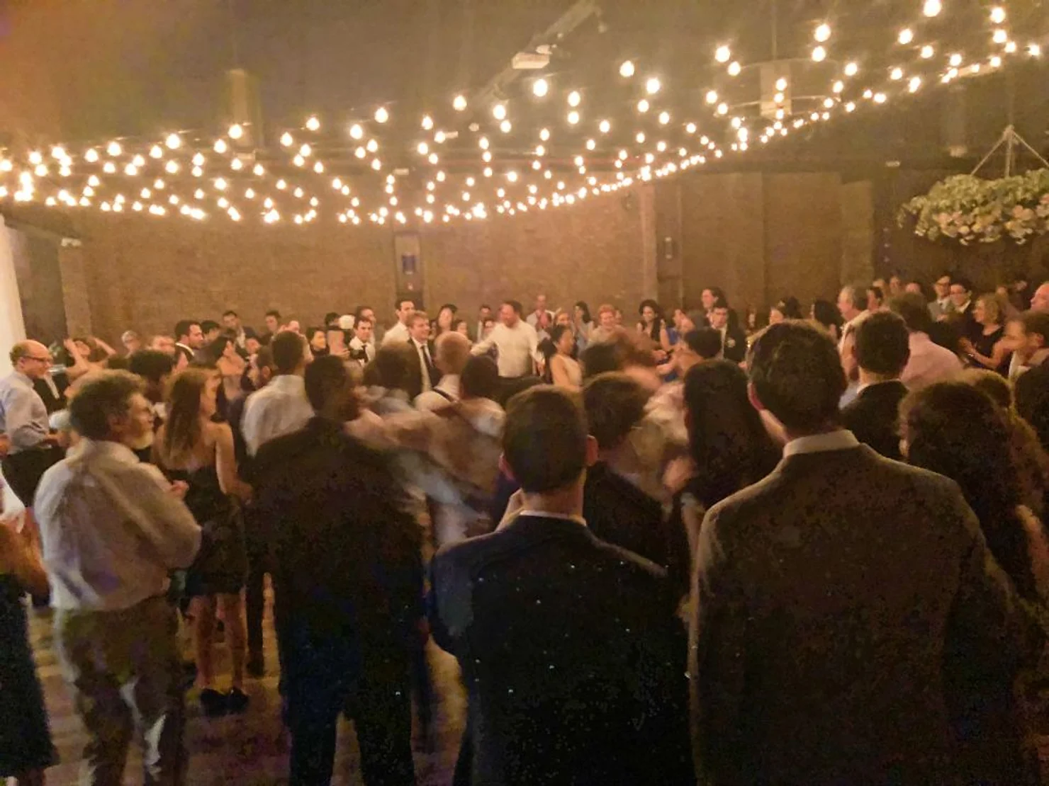 NYC 26 Bridge Wedding DJ Crowded wedding dance floor under warm string lights in a hall, guests dancing and mingling in a lively celebration.