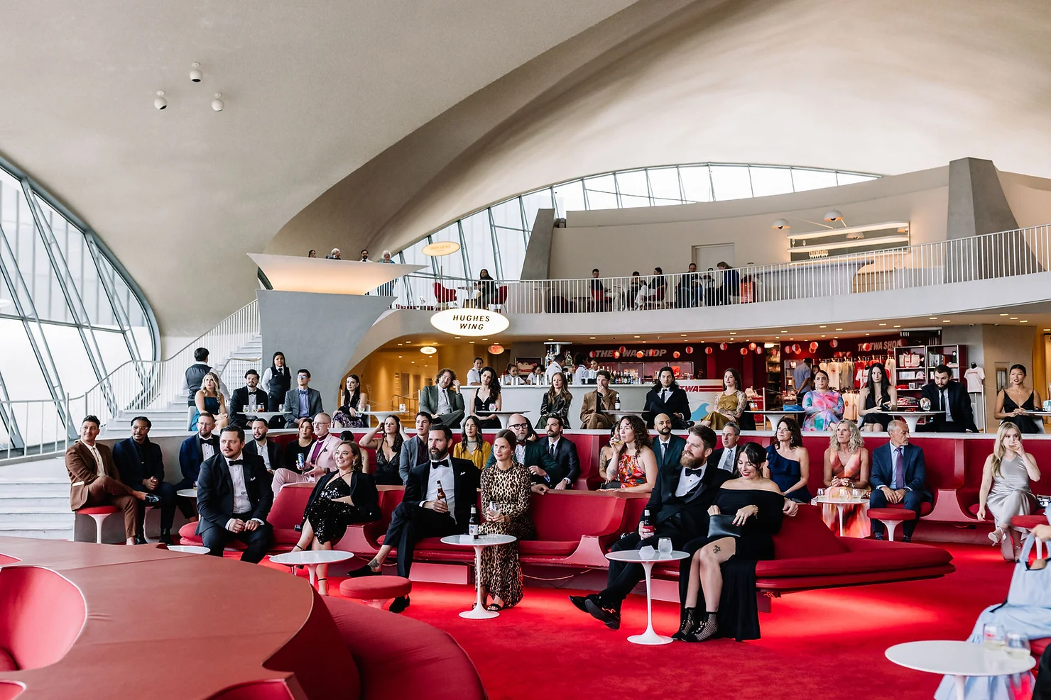 nyc non traditional wedding dj Crowd of formally dressed adults in a modern lounge with red seating under a curved ceiling, watching an event near Hughes Wing signs.