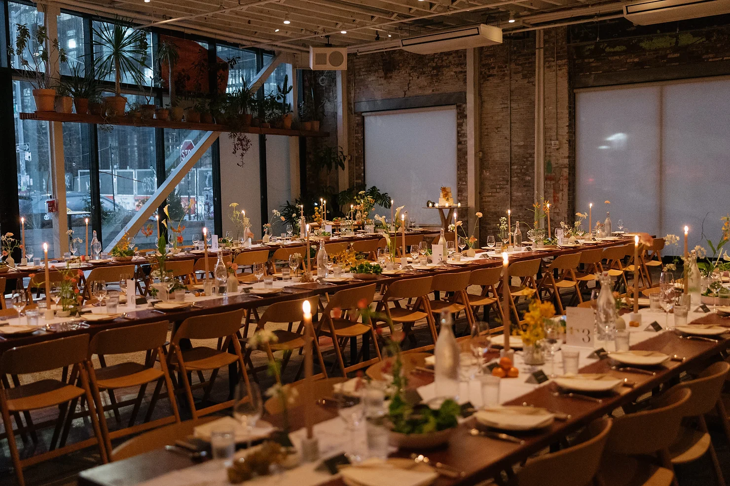 nyc rule of thirds wedding dj. Candlelit banquet tables set in a plant-filled industrial room with brown chairs, glassware, and warm evening glow