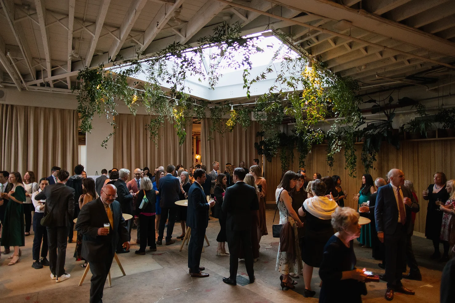 nyc rule of thirds wedding dj . Guests mingle at a formal indoor reception under hanging greenery and skylights in a loft-like hall.