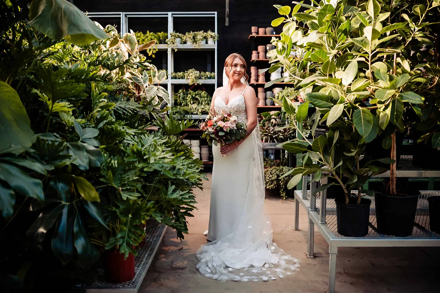 Terrain Gardens at DelVal in Doylestown wedding dj Bride in white gown holding a bouquet stands in a lush plant nursery, surrounded by green foliage and shelves of potted plants.
