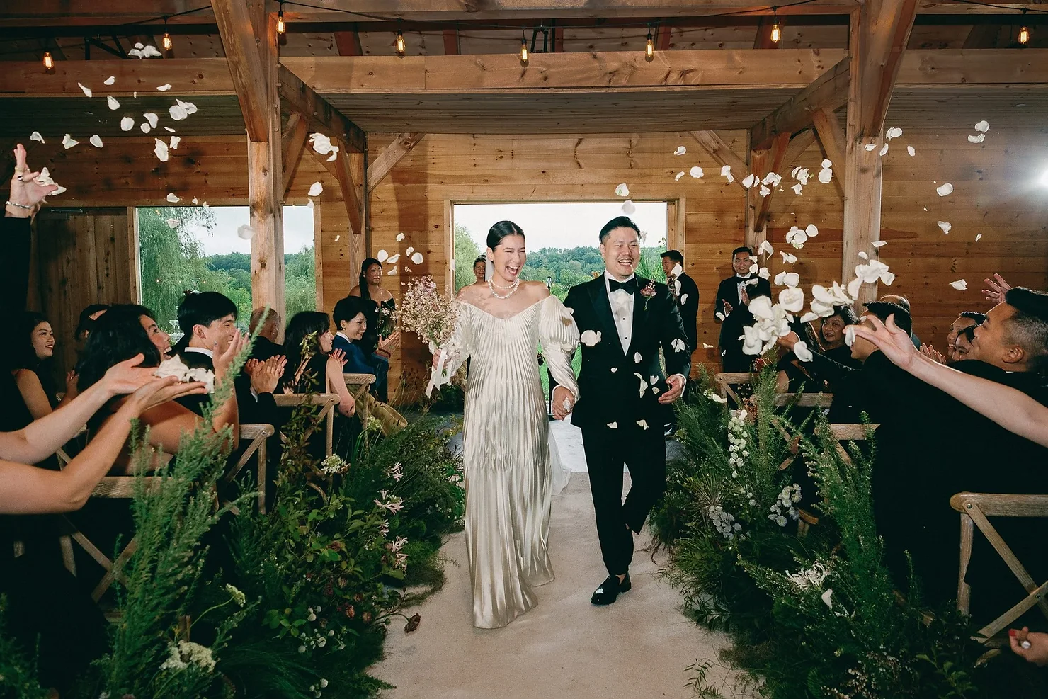 nyc ulster county wedding dj Newlyweds in white gown and black tux walk down a wooden barn aisle as guests toss flower petals.