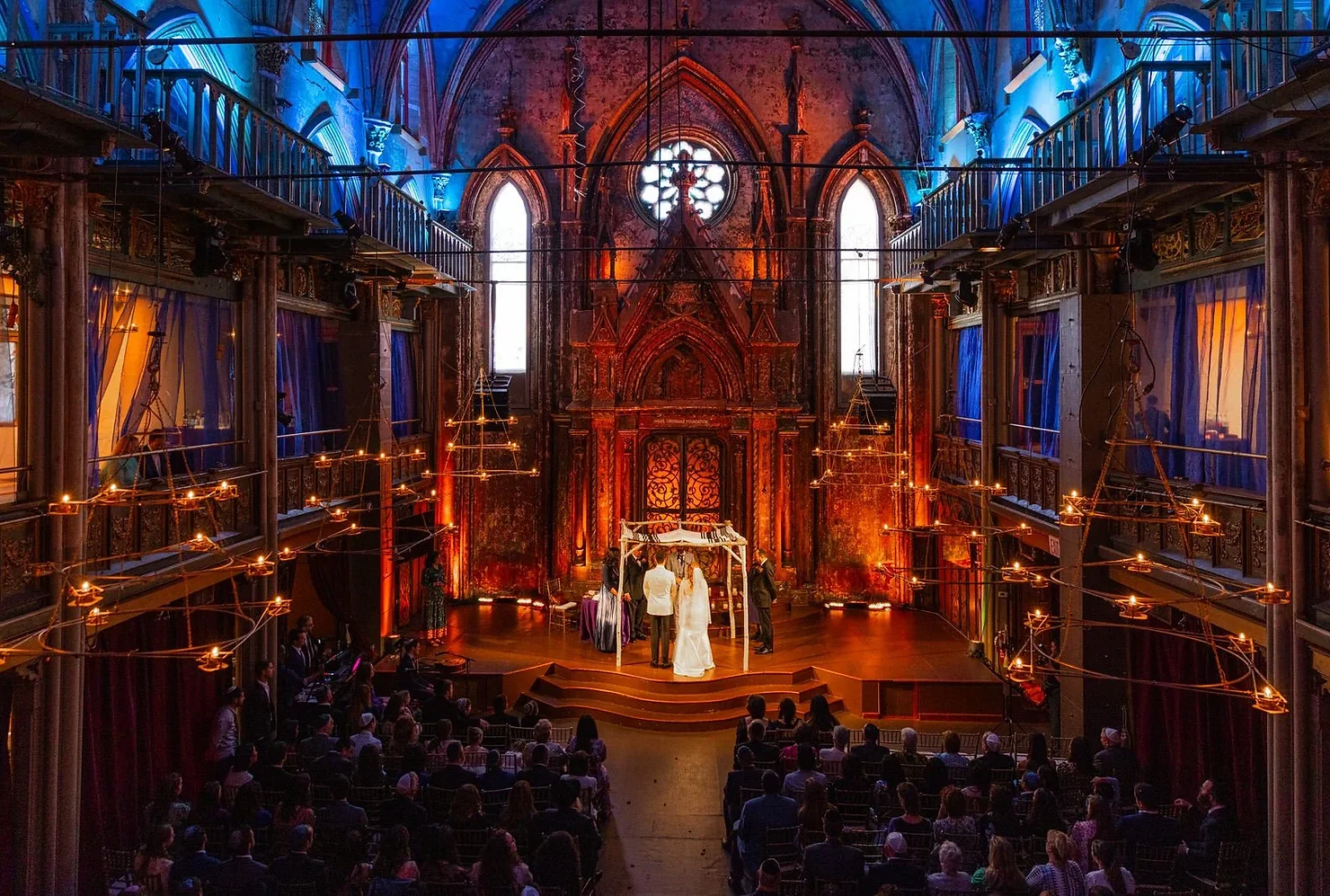 nyc non traditional wedding dj Wedding ceremony on a stage in a Gothic hall, couple at the altar before seated guests under blue and orange lights.