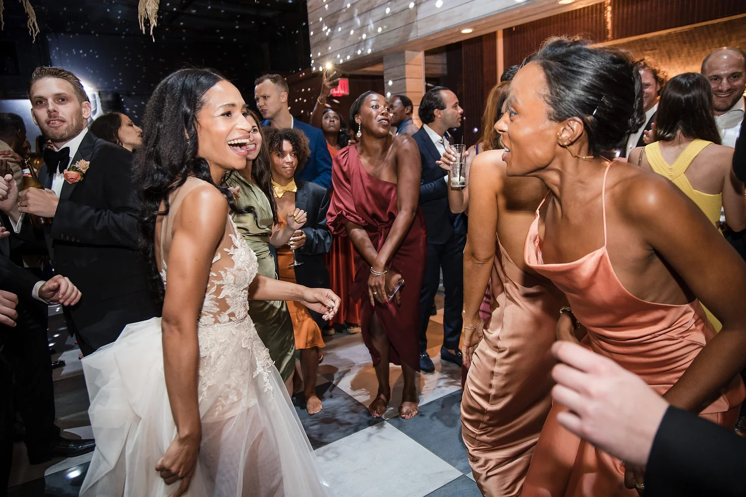 nyc weddind dj wythe hotel Wedding guests dance and laugh at a lively reception, with a bride in a lace gown and women in satin dresses in a dark, modern venue.