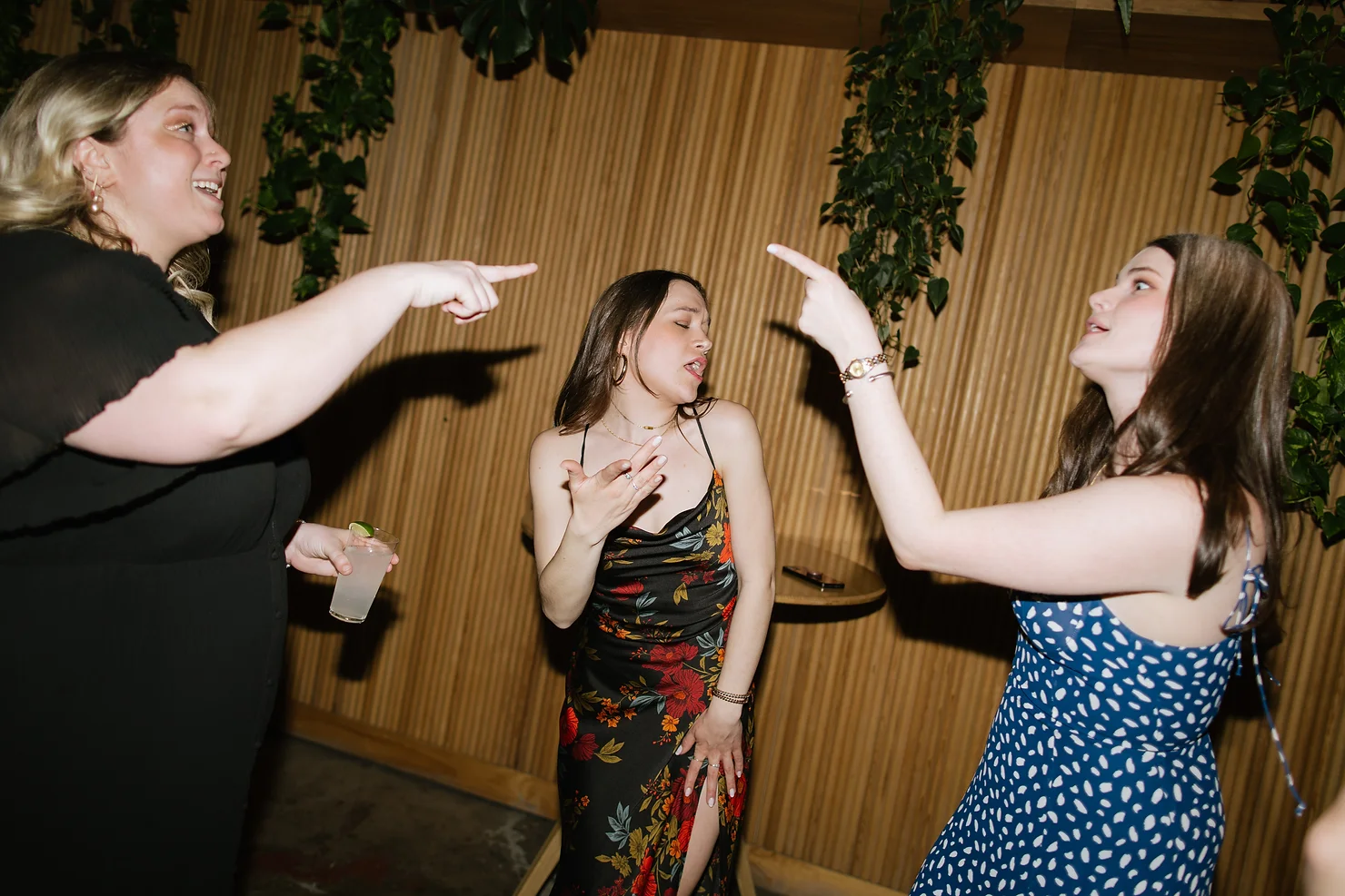 nyc wedding dj Three women at a party point at each other and talk animatedly; one holds a drink, in front of a wood wall with hanging greenery.