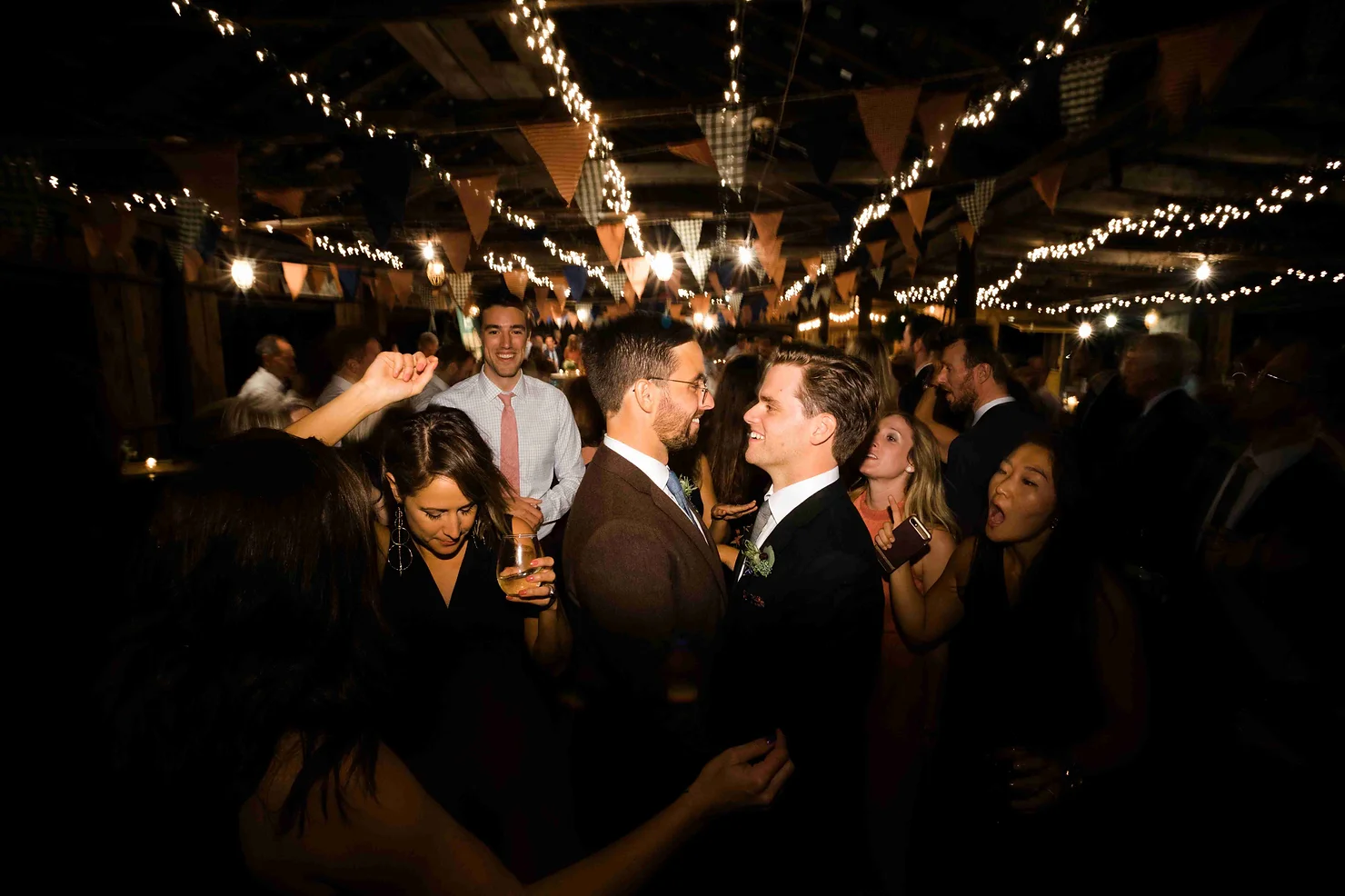 non traditional wedding dj catskills for gay weddings: Guests dance and mingle at a warmly lit indoor party under string lights and bunting, with smiling couples in suits and dresses.