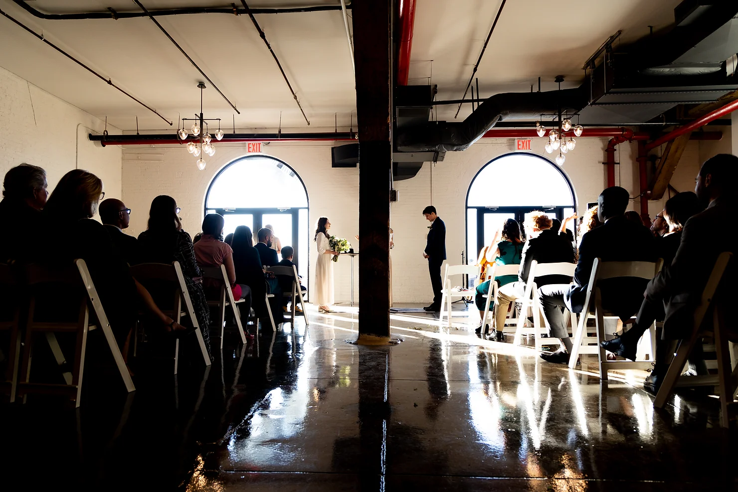 NYC wedding DJ Strong Rope Brewery Bride and groom stand before guests in a bright industrial loft wedding ceremony, with EXIT signs and polished concrete floor.