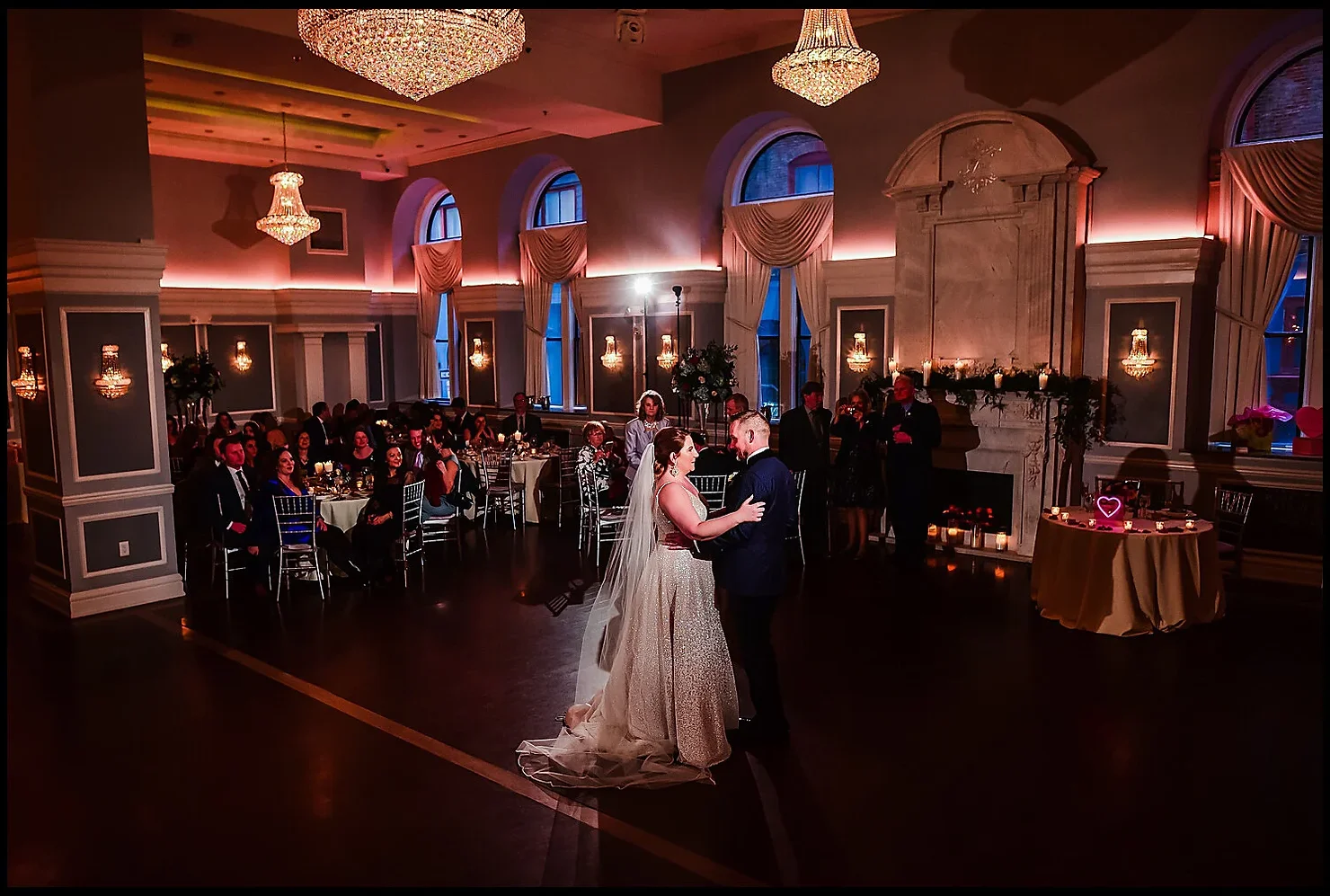 Philly wedding dj Bride and groom slow-dance at a wedding reception in an elegant chandelier-lit hall, with seated guests watching nearby.