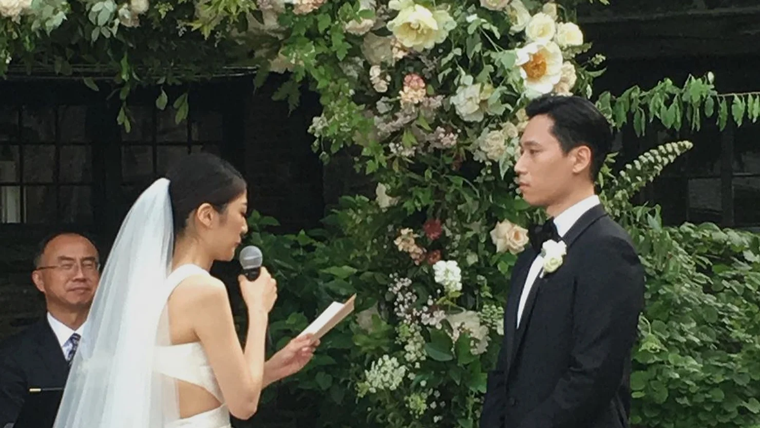 nyc Blue Hill at Stone Barns non traditional wedding dj: Bride reads vows into a microphone to the groom at an outdoor wedding beneath a flower arch, while the officiant watches.