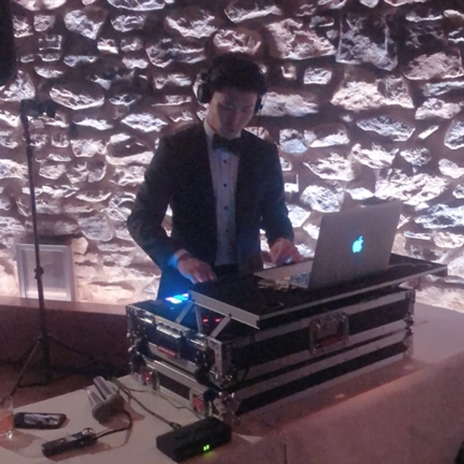 nyc wedding dj Man in a tux DJing at a laptop and mixer, against a lit stone wall; Apple logo glows on the laptop.