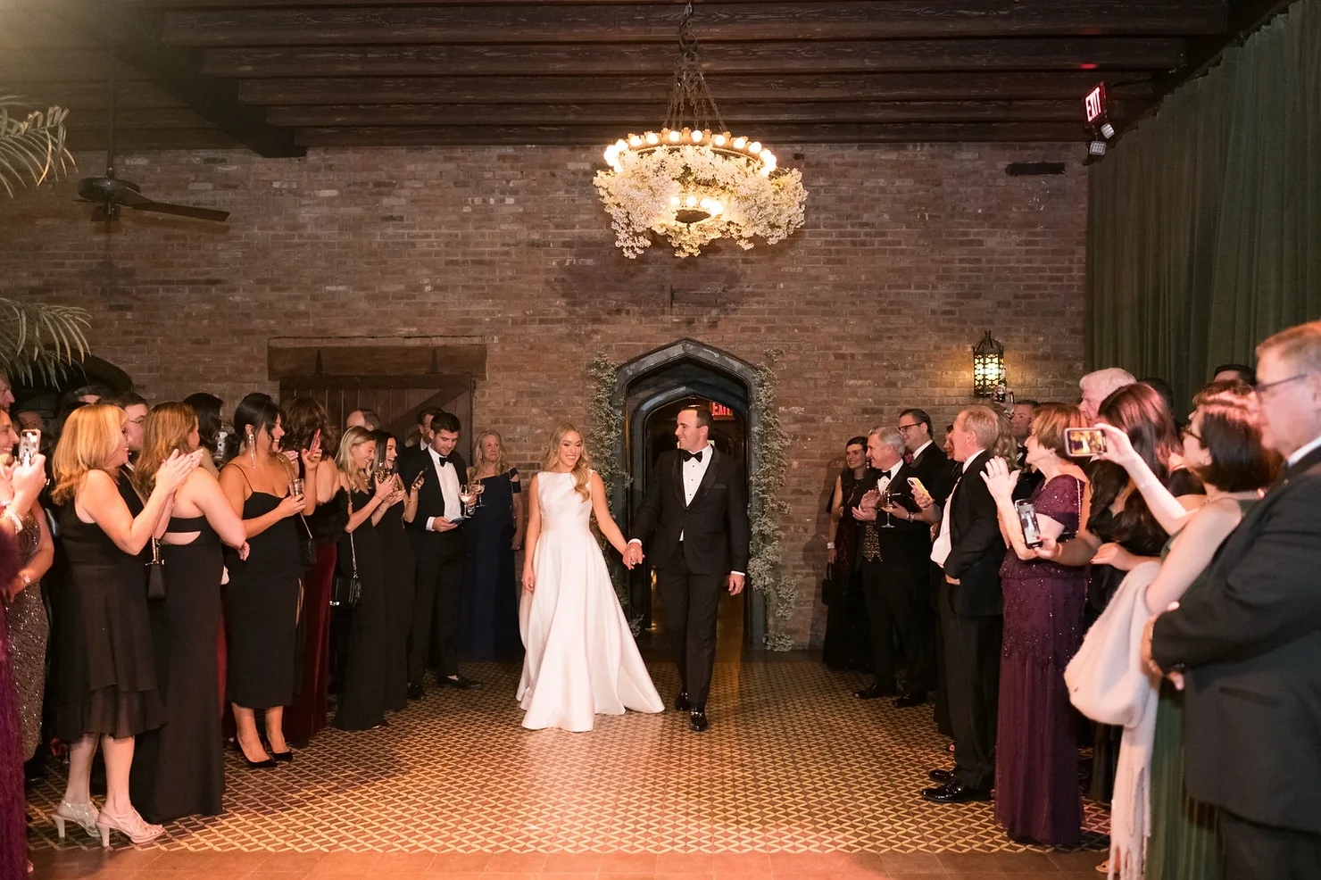 The Bowery Hotel NYC Wedding DJ Bride in white gown and groom in black tuxedo walk hand in hand through applauding guests in a brick-walled hall.