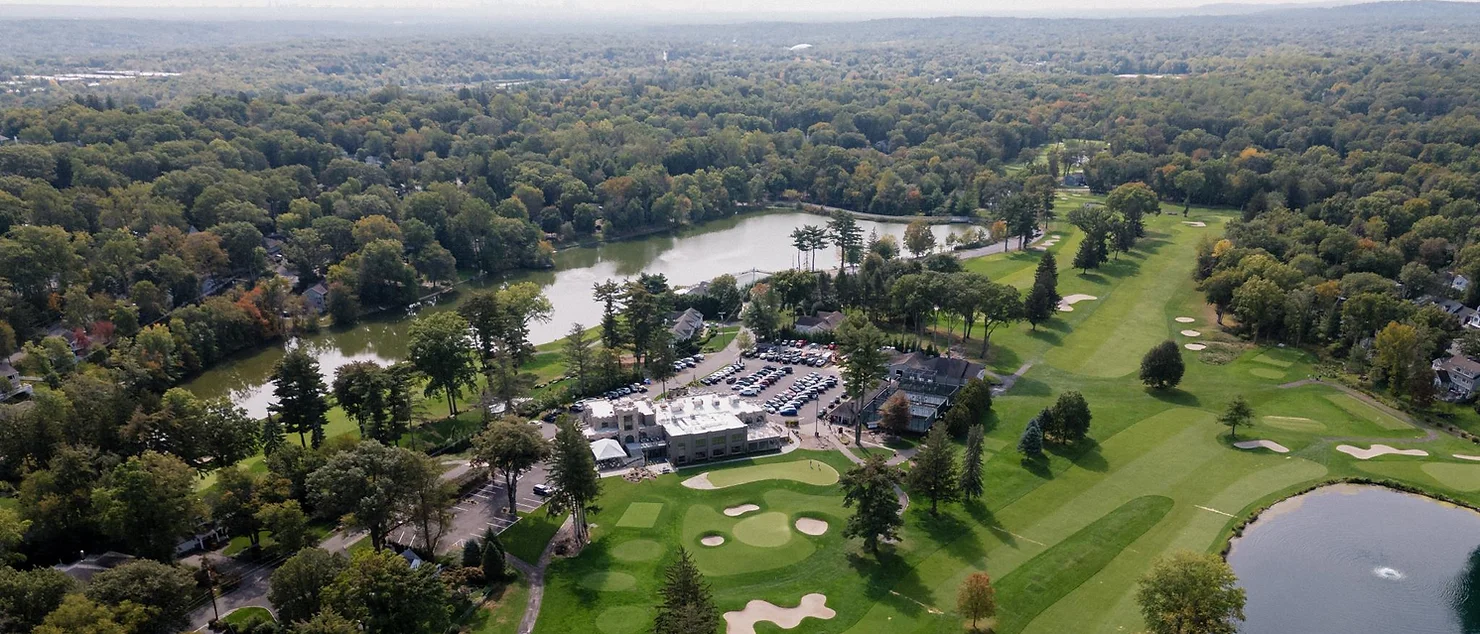nyc wedding dj bergen county dj Aerial view of a golf course with sand traps, clubhouse, parking lot, and lake surrounded by dense forest.