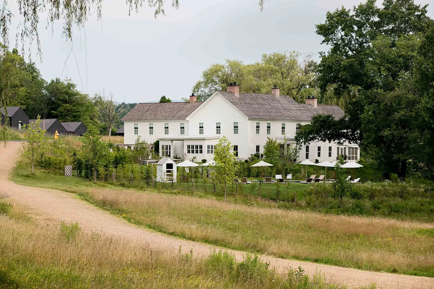 nyc ulster county wedding dj Large white farmhouse with lawn umbrellas beside a gravel path, surrounded by green trees and grassy fields under a calm sky