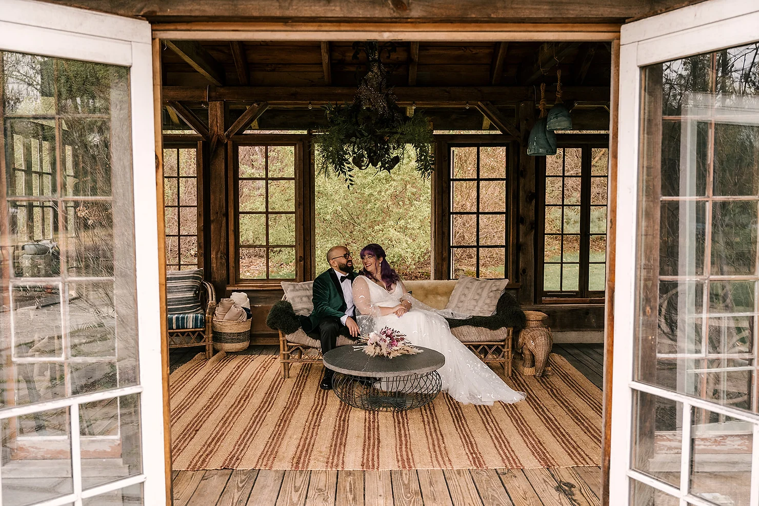 Foxfire Mountain House Wedding DJ: Smiling bride and groom sit on a rustic porch sofa in a wooden pavilion, with windows, woven rug, and floral decor.