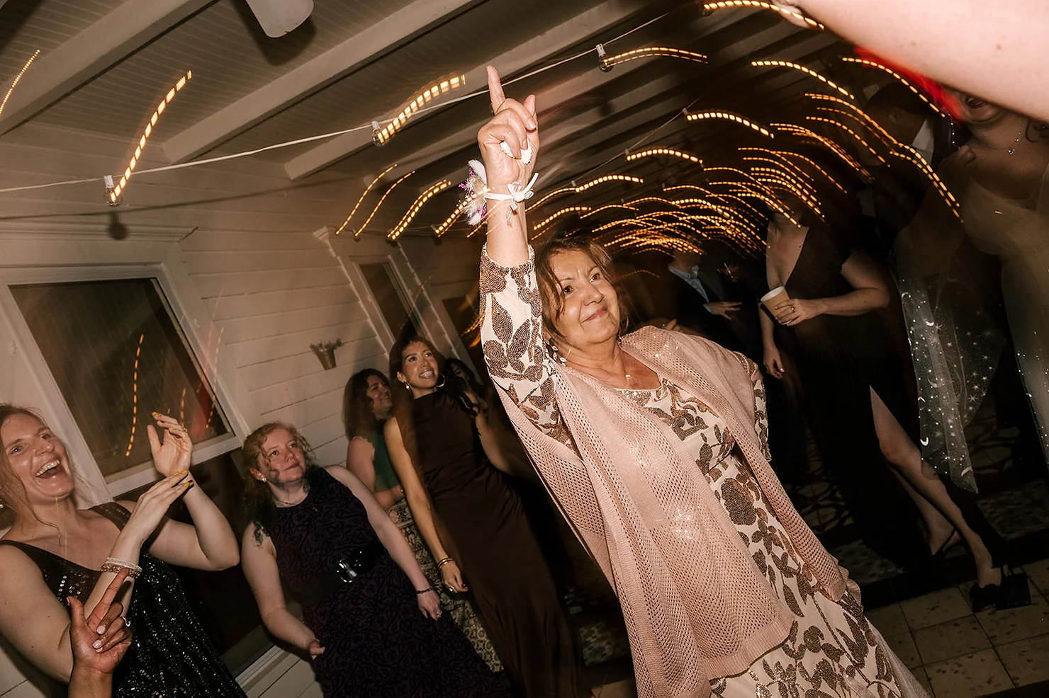 nyc wedding dj foxfire mountain house Woman in floral dress and pink shawl points upward at a lively party, surrounded by dancing guests under warm string lights.