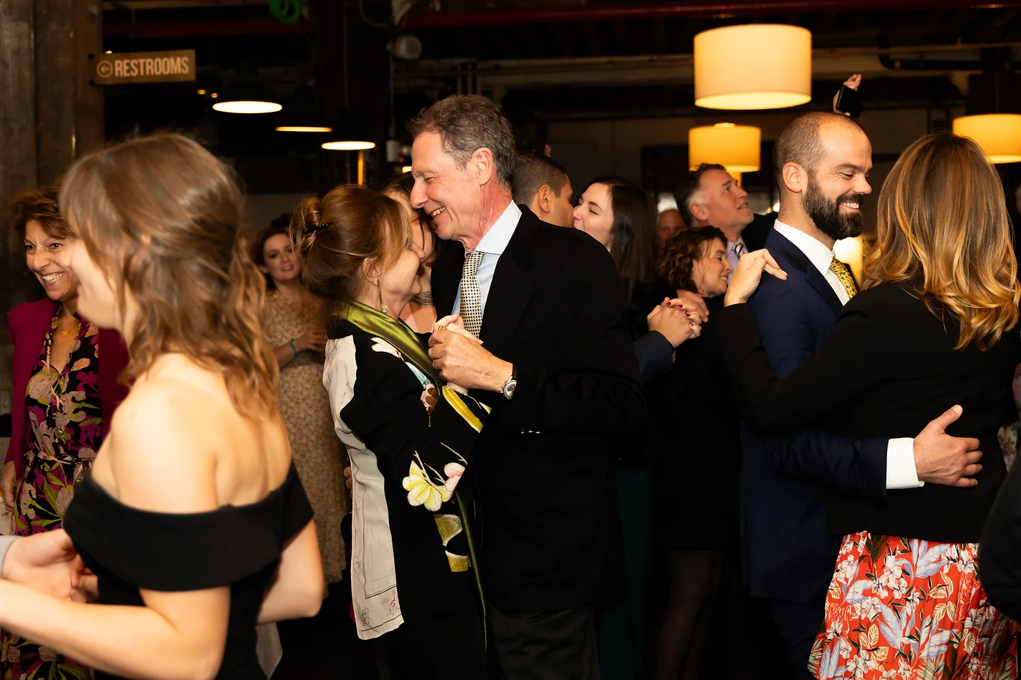 NYC wedding DJ Strong Rope Brewery Partygoers dance in pairs under warm lights at an indoor event, with a Restrooms sign overhead and smiling faces.