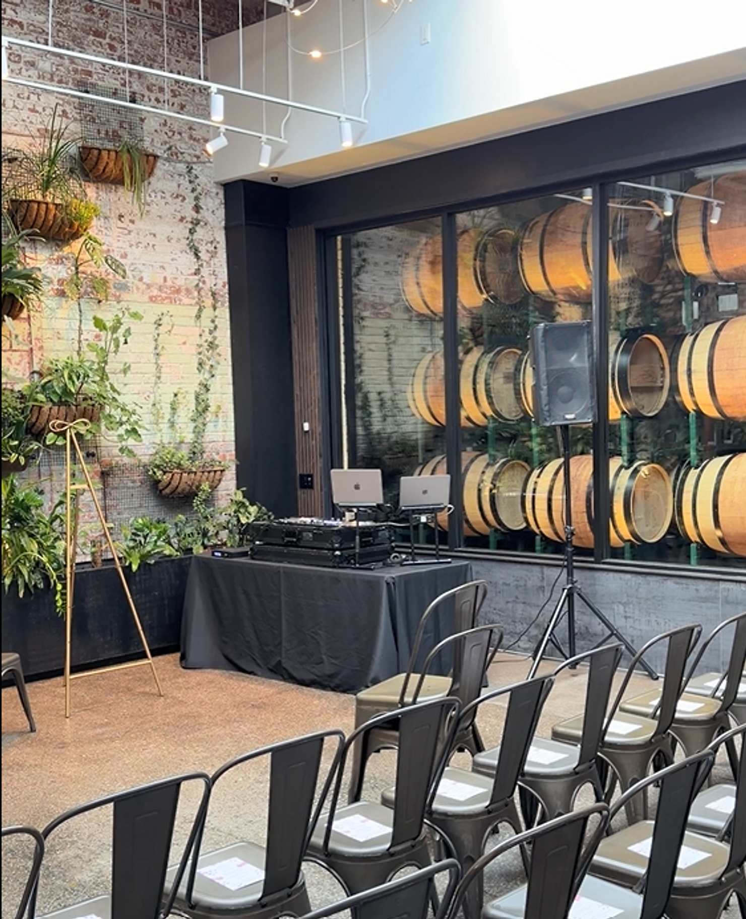 nyc non traditional wedding dj brooklyn winery Empty event venue with rows of chairs, a DJ table, and warm-lit wine barrels behind glass beside a plant wall.