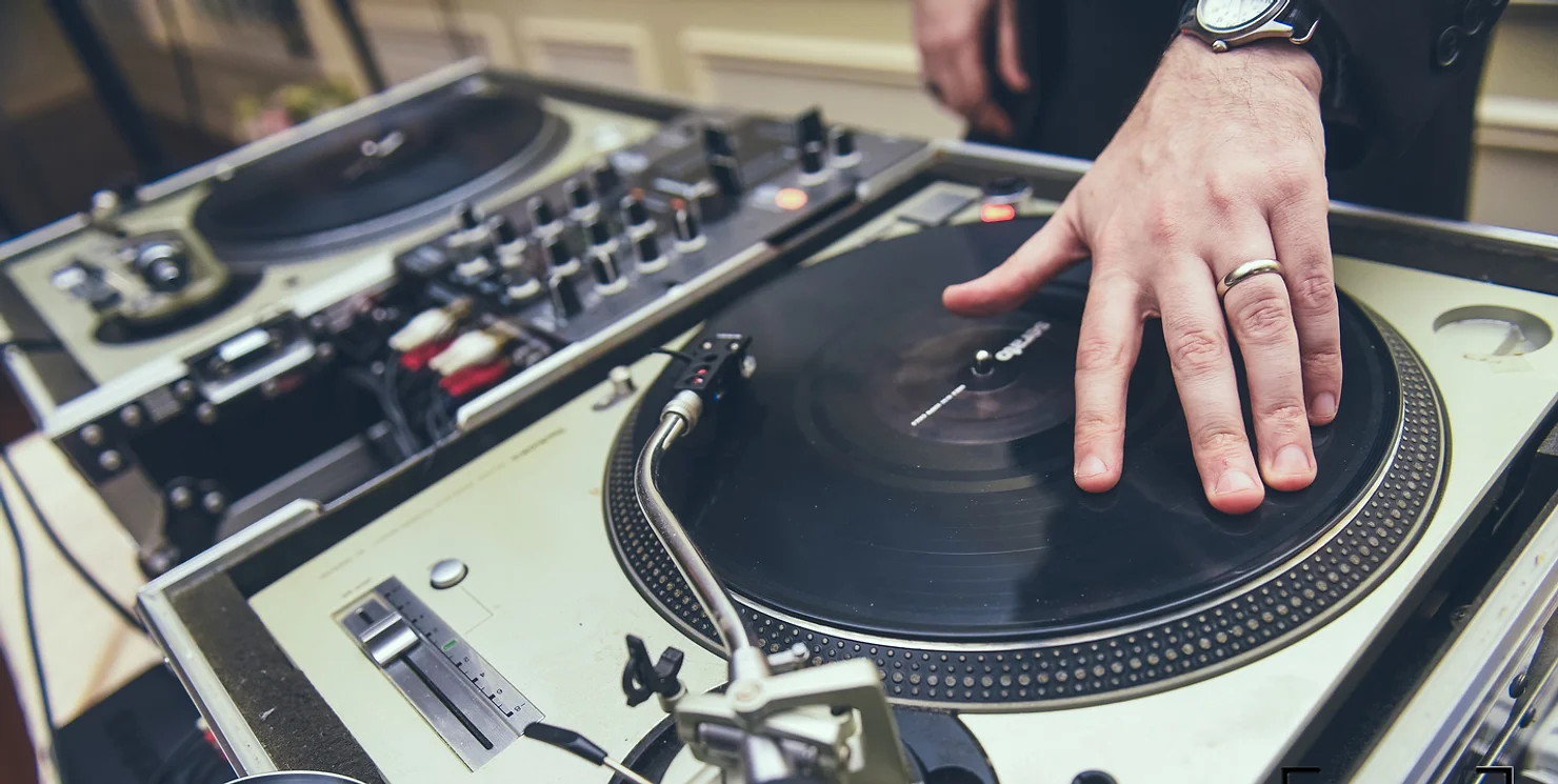 Long Island Wedding DJ A DJ's hand with a ring spins a vinyl record on a turntable with DJ equipment in the background, suggesting a lively music scene.