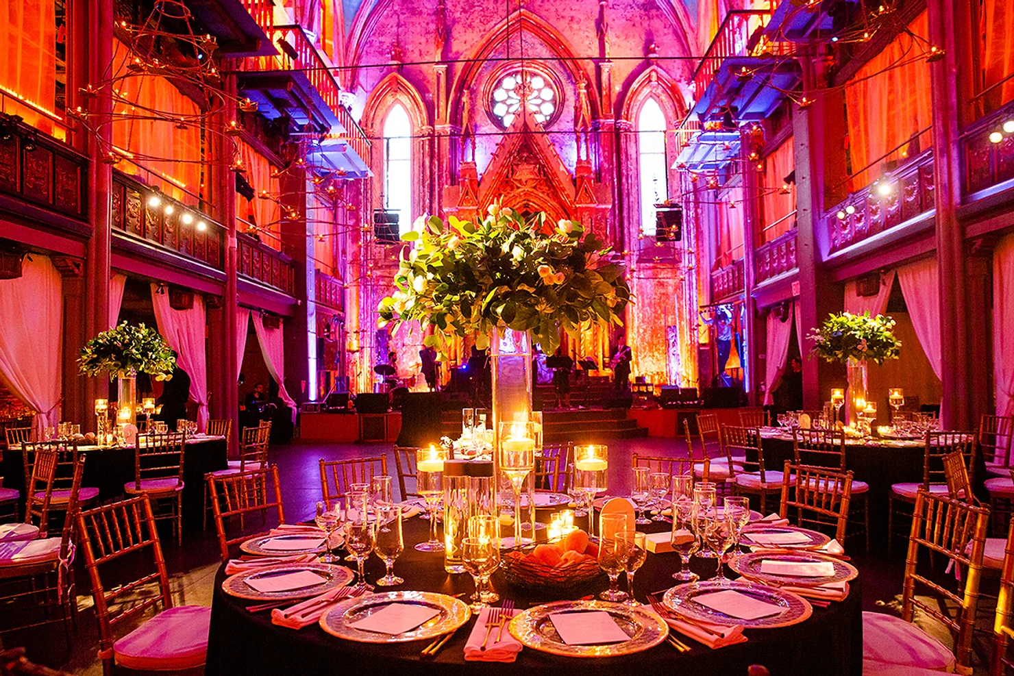 NYC wedding DJ Angel Orensanz Foundation Candlelit banquet in a grand  hall, with round set tables, floral centerpieces, and vivid pink-purple lighting on stage.