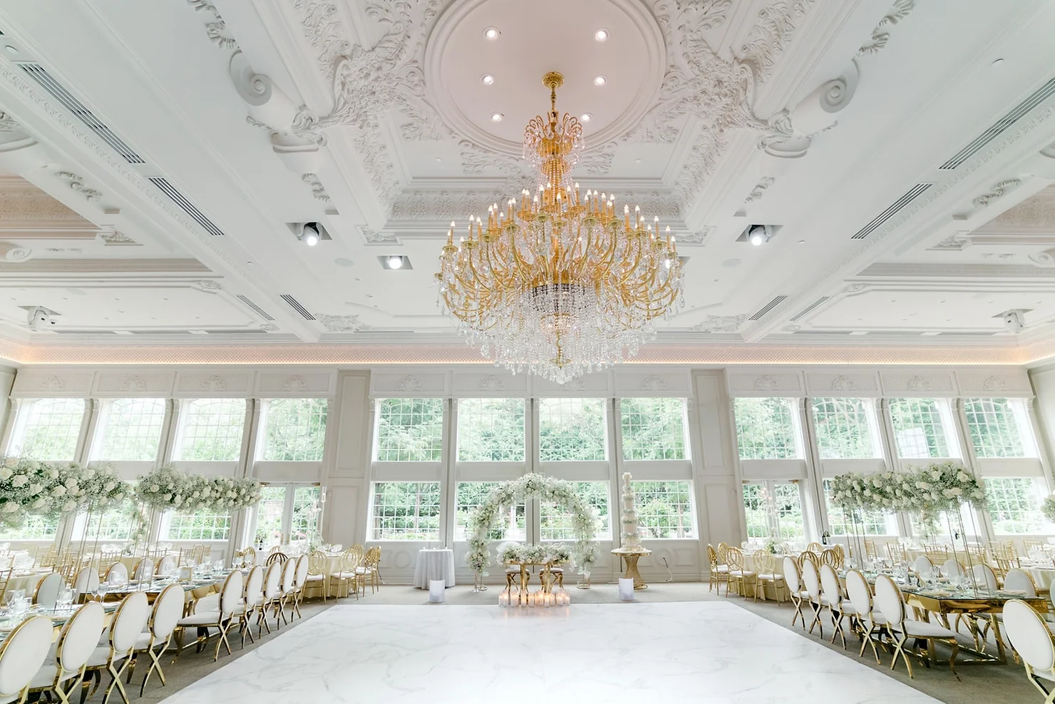 nyc wedding dj bergen county dj Elegant white ballroom with gold chandelier, floral arch, and set dining tables for a wedding reception; no people present