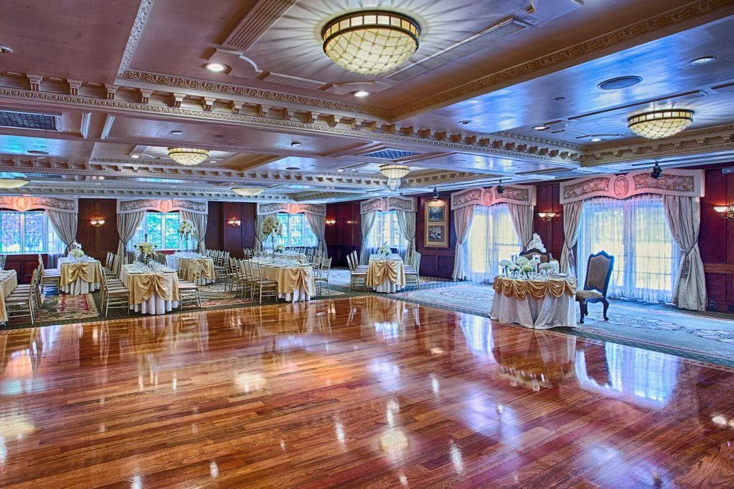nassau county nyc wedding dj Elegant empty banquet hall with polished wood floor, set round tables, chandeliers, draped windows, and soft pink-blue lighting