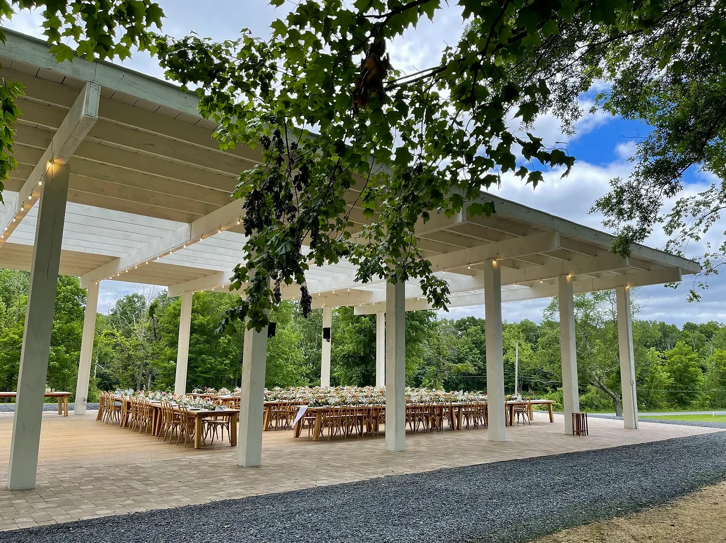 nyc wedding dj foxfire mountain house Open-air pavilion set for an outdoor event, with long wooden tables and chairs beneath leafy trees and a blue sky