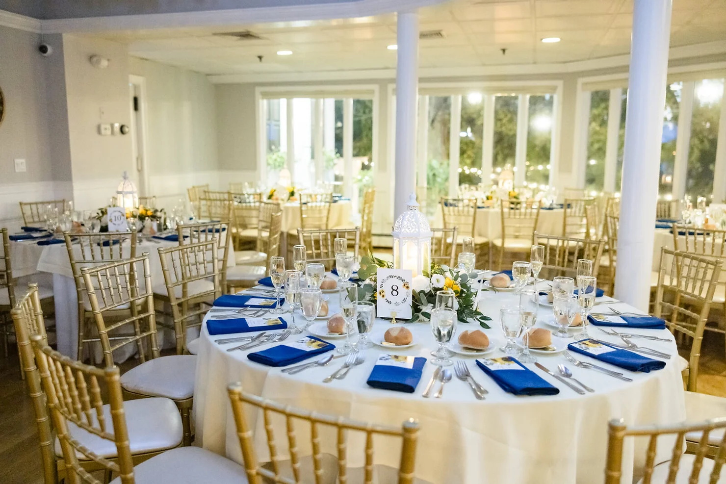 nassau county nyc wedding dj Elegant banquet hall with round tables set for a reception, blue napkins, gold chairs, lantern centerpieces, table 8 sign.