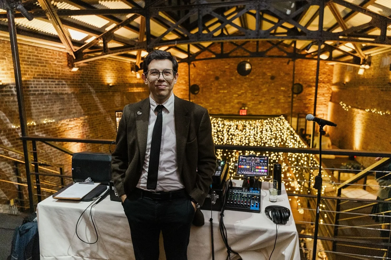 NYC Wedding DJ in suit stands by audio mixer and microphone in a brick loft venue lit with warm string lights.
