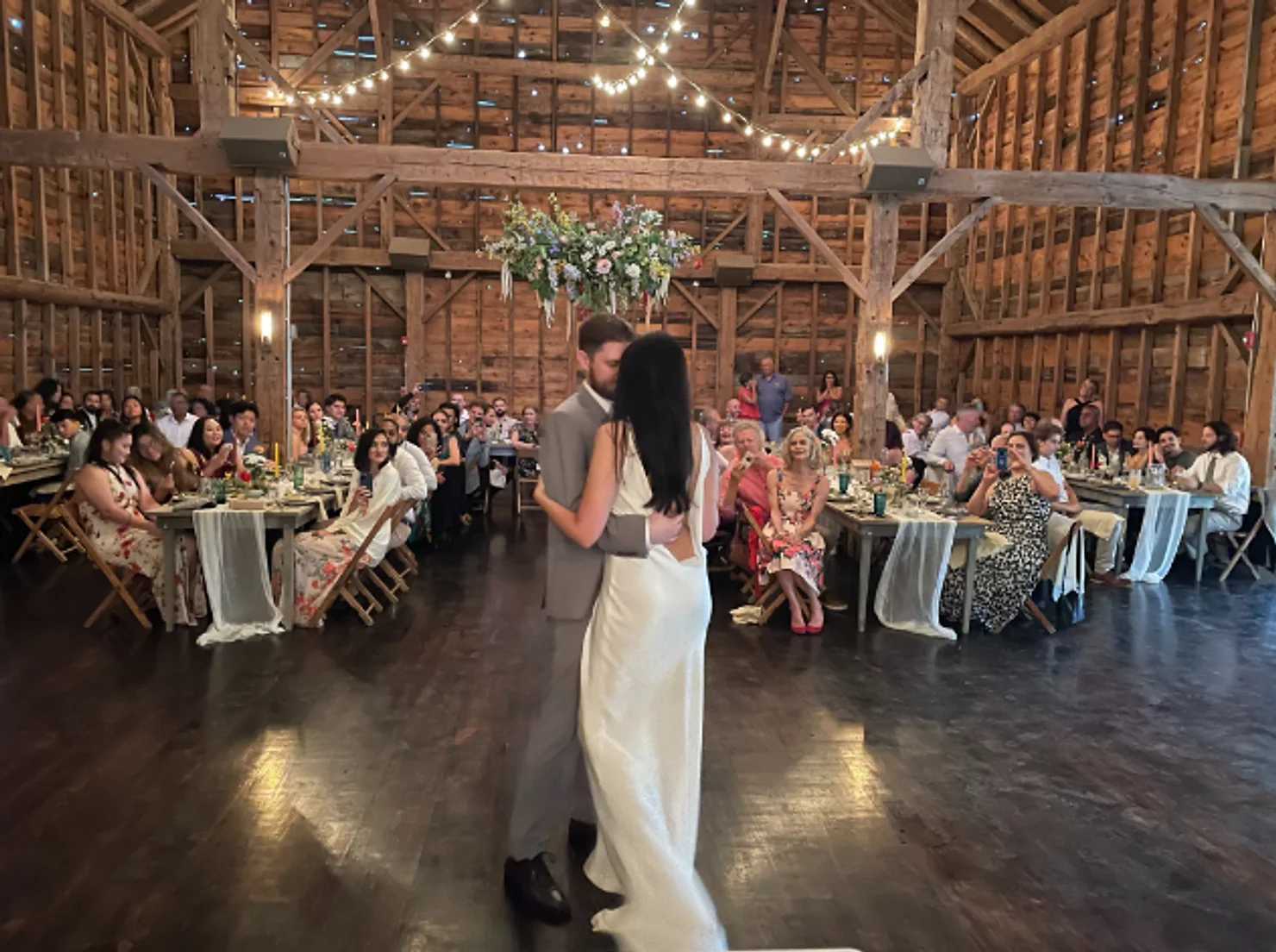 nyc catskills wedding dj Bride and groom slow dance in a rustic barn reception, guests seated at candlelit tables under string lights.
